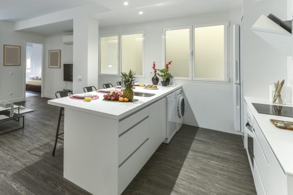 A kitchen island with a white countertop, cabinets with drawers, black stools and a breakfast service with a lot of assorted fruit and orange juice in glasses